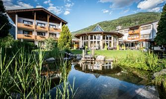 An idyllic hotel grounds with an adjacent pond and plenty of greenery. Behind the pond are the hotel's buildings, surrounded by mountains and blue skies.