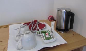 A tea station on a wooden shelf with two cups, spoons, and a kettle. Next to it are sugar and tea bags in a small bowl.