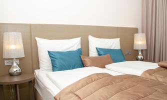 A cozy bedroom with two beds, white bed linens, and decorative blue pillows. Next to the beds are elegant nightstands with lamps.