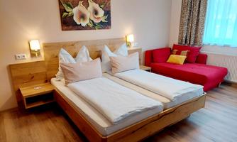 A cozy bedroom with two twin beds and wooden decor. The room features a red sofa and a flower painting on the wall.