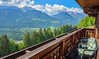 A beautiful view from a balcony overlooking green hills and mountains under a clear blue sky. Two chairs are ready to enjoy the landscape.