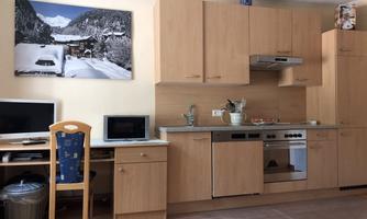 A modern kitchen with wooden cabinets and appliances. A desk with a computer and a picture of snow-covered mountains are also visible.