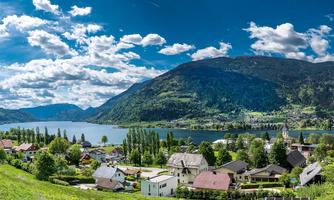 A picturesque landscape with a clear lake, surrounded by mountains and green forests. Small houses are located near the water under a bright blue sky.