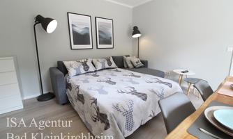 A modern bedroom with a large bed and cozy sofa. The walls are painted light and adorned with artworks, while a table and chairs stand in the foreground.
