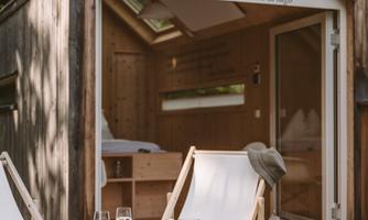 A cozy terrace in front of a modern wooden house by the water. On a table there are drinks and snacks, surrounded by relaxed lounge chairs.