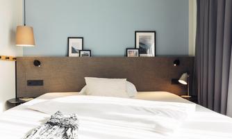 A modern bedroom with a large bed and a light wall color. There are pictures hanging on the wall, and there is a lamp as well as curtains.