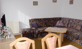 A cozy living space with a large, patterned sofa and a table in the center. Pictures hang on the walls, and there is a floral decoration on the table.