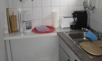 A small, functional kitchen with a sink and modern appliances. The walls are brightly tiled, and there is a wooden shelf with kitchen utensils.