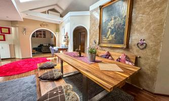 An inviting dining room with a large wooden table and cozy seating cushions. A classical painting hangs on the wall, and additional living areas are visible in the background.