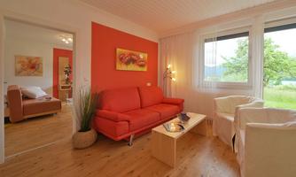 A cozy living room with a red couch and two white chairs. Large windows offer a view of the nature outside.