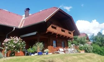 A traditional wooden house with a red roof, surrounded by a well-tended garden. The sunny landscape and the blue sky give the scene an inviting atmosphere.