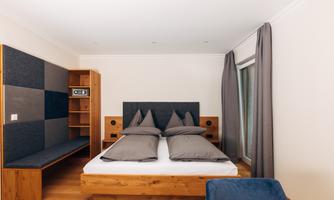 A modern bedroom with a wooden bed and gray cushions. The walls are painted in light shades, and there is a cozy seating area with a shelf.