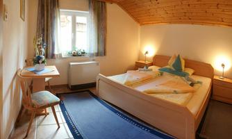 A cozy room with a double bed, a desk, and a window. The wooden walls and soft light create an inviting atmosphere.