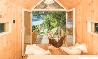 A cozy, bright room with wooden paneling and large windows. The view overlooks a green landscape and a wooden walkway.