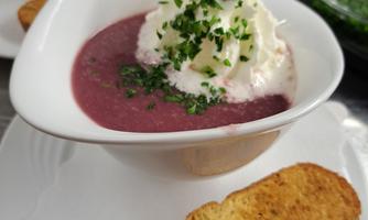 A creamy soup in a white bowl, garnished with cream and fresh herbs. Next to the bowl is a crunchy piece of bread.