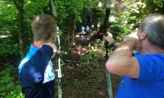 Two men are standing in the forest, aiming with bows at targets. The surroundings are green and natural.