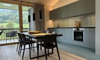 A modern kitchen with a dining table and chairs. Large windows provide a view of the green landscape.