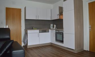 A modern kitchen with white cabinets and wooden details. The floor is made of wood, and across from it is a black couch.