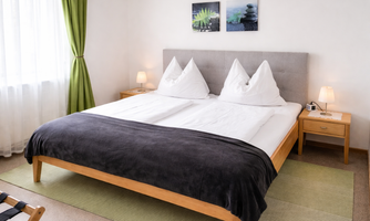 A cozy bedroom with a large double bed and clean comforters. The room is bright and cheerful, with green curtains and modern bedside lamps.