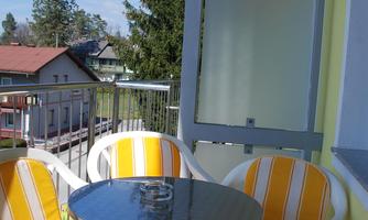 A cozy balcony with a round table and four white chairs that are upholstered with yellow and white stripes. In the background, trees and other houses are visible.