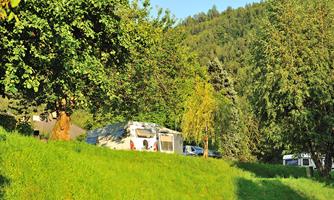 A caravan is situated on a green meadow surrounded by trees. In the background, gentle hills can be seen.