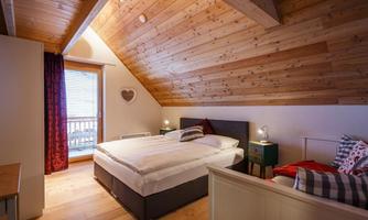 A cozy bedroom with wooden walls and a sloped roof. It contains a large bed, nightstands, and a seating area with a window to the outside.