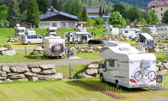 A campsite with several RVs in a green environment. Trees and buildings can be seen in the background.