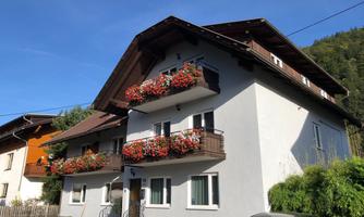 A charming house with balcony flowers and a clear blue sky. The surroundings show a calm, rural atmosphere.