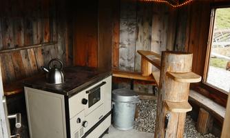 A rustic room with wooden walls and an old stove. In the corner stands a teapot and there is a small seating area made of wood.