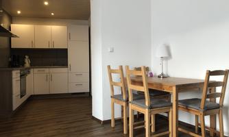 A modern kitchen with white furniture and a wooden dining table. The room has a cozy atmosphere and warm wooden floor.