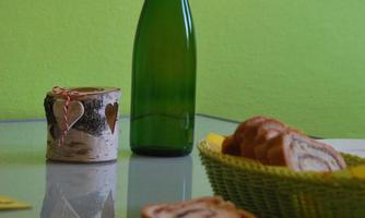 A table with a green wall in the background. On it are a bottle, a glass with hearts, and a bowl with bread.