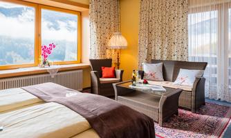 A cozy hotel room with two beds and a living area. Large windows offer a view of the mountains and plenty of daylight.