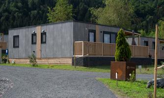 A modern wooden house with a spacious veranda, surrounded by a green landscape. The path leads to the entrance of the house and is covered with gravel.