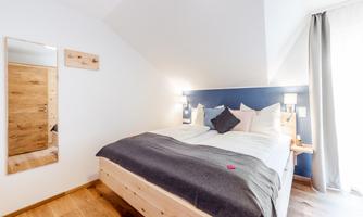 A modern bedroom with a large bed and natural wooden furniture. The walls are bright and the windows let in a lot of light.