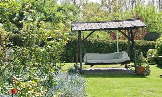 A beautiful garden with a wooden swing under a roof. Surrounded by colorful flowers and lush greenery.