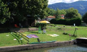 A peaceful landscape area by the water with green meadows and lounge chairs. In the background, trees and a blue sky can be seen.