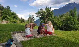 A group of children is playing outside and exploring a sandbox. In the background, green trees and mountains are visible.