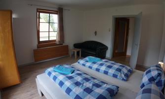 A bright room with two single beds covered with checked blankets. There is a sofa, a table, and a large window that lets in light.