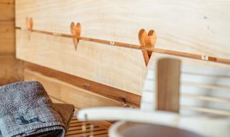 A wooden chair in a sauna with a towel and a wooden bowl. The walls are made of wood and have heart-shaped cutouts.