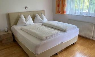 A cozy bedroom with a large double bed and two pillows. The room has wooden flooring and large windows with curtains.
