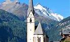 A beautiful church with a tall tower in a mountainous landscape. In the background, snow-capped mountains and a blue sky can be seen.