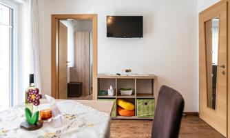 A cozy room with a table, a television on the wall, and a shelving unit with colorful storage boxes. The decor is modern and bright.