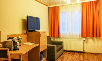 A simple room with a television, a desk, and a comfortable armchair. The walls are light, and the orange curtain lets in daylight.