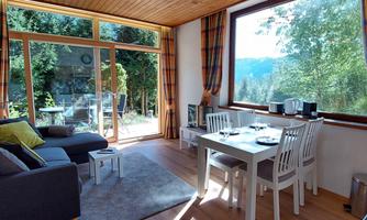 A bright and cozy living room with a dining table and a sofa. Through large windows, one looks out onto a green landscape.