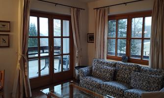 A cozy living room with a blue sofa and a glass table. The windows offer a view of the terrace and the outdoor area.