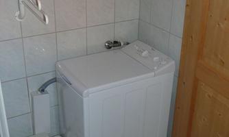 A small laundry room with a washing machine and a white stool. The walls are tiled and the room has a wooden door.
