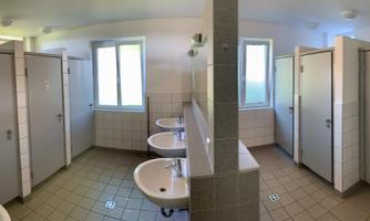 A bright bathroom with multiple sinks and closed cabin doors. There are windows that let in daylight.