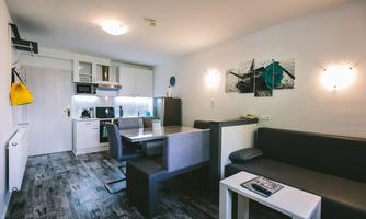 A modern kitchen unit in a bright living space with a dining table and a cozy seating area. The walls are tastefully decorated and the floor features an elegant design.