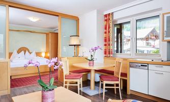 A bright, modern room with a living area and a kitchenette. In the foreground, there is a table with a flower arrangement and chairs.