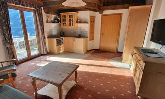A cozy living space with wooden furniture and a large window that lets in natural light. In the background, there is a small kitchen and a television visible.